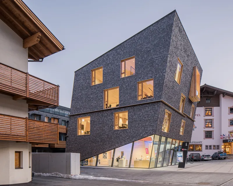 Three-story shingled volume with illuminated windows and an angled glass storefront at dusk