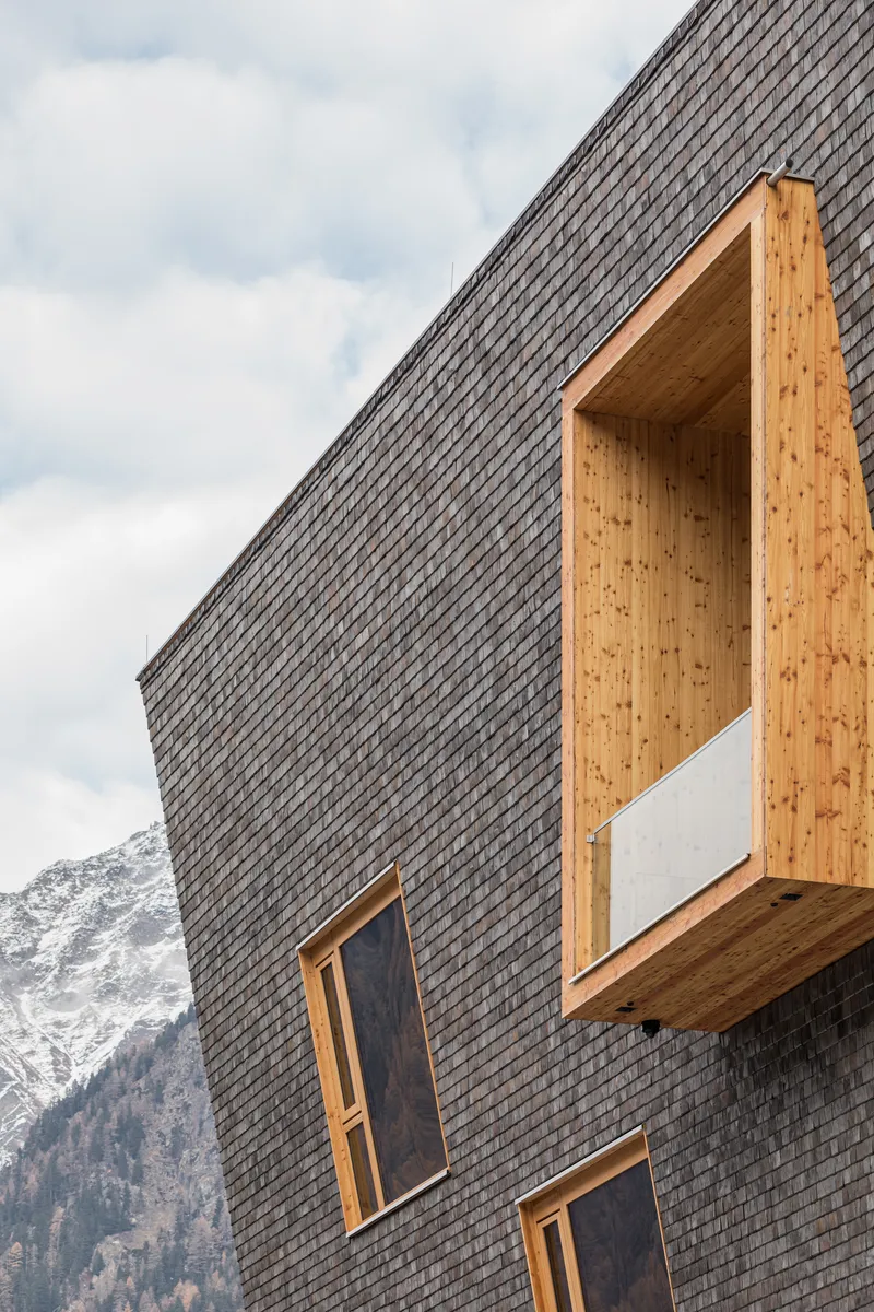 Close-up of a cantilevered timber balcony projecting from the dark shingled facade with mountains behind