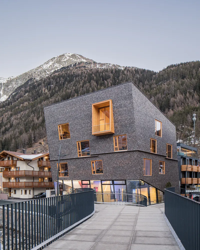 Approach to the shingled building from a pedestrian bridge with forested mountain slopes behind