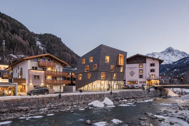 Cubic volume clad in dark shingles nestled between traditional alpine buildings along a winter stream