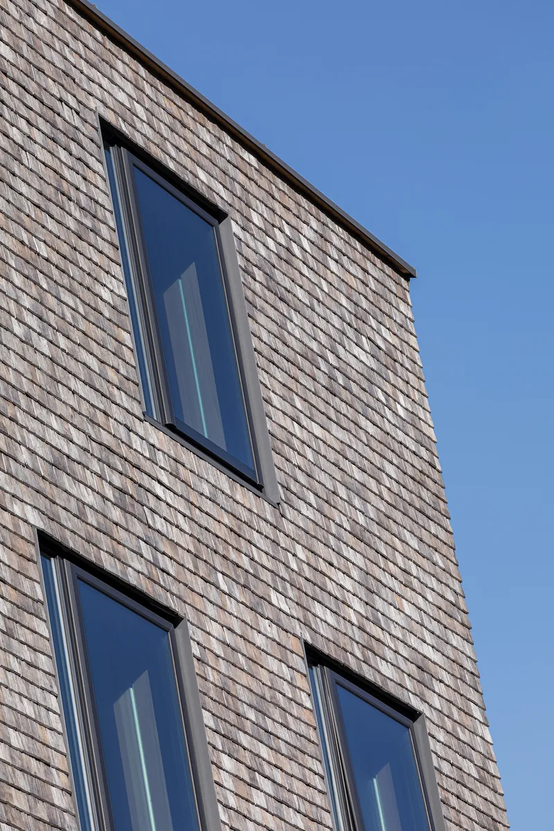 Close-up of three angled windows punched into a pale grey shingle facade against blue sky