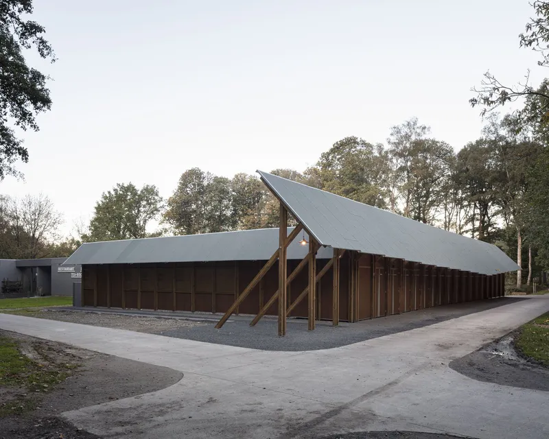 Long pavilion with timber portal frames and metal roofing beside a paved path on an overcast autumn day