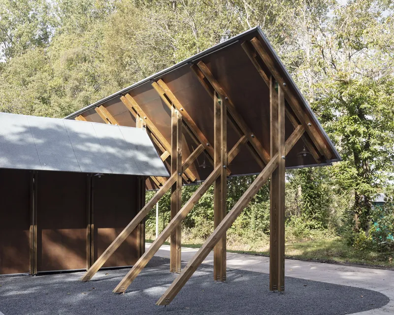Exposed timber portal frame with diagonal bracing forming an open entrance canopy in dappled sunlight