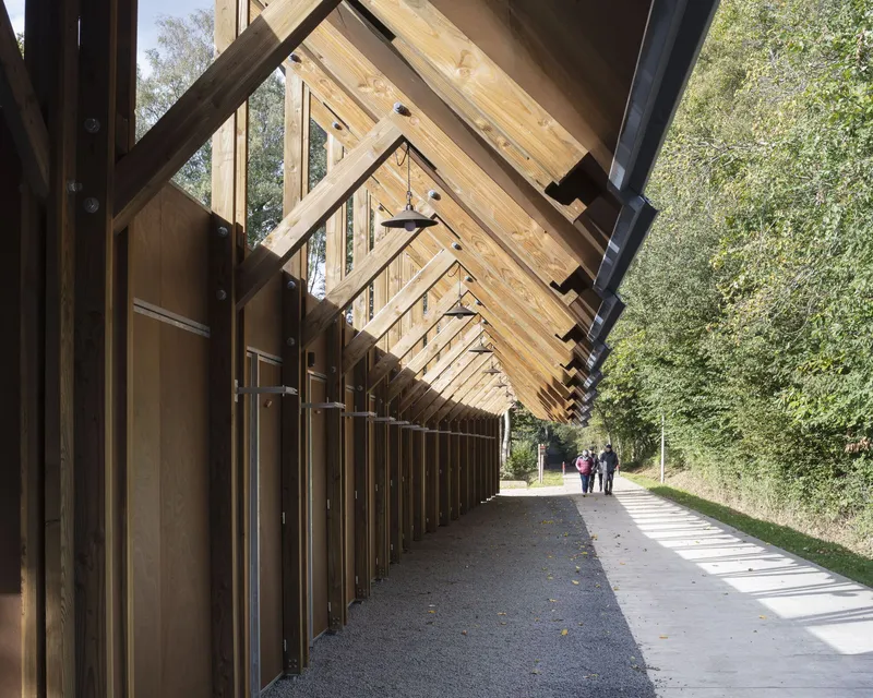 Covered walkway with sawtooth timber rafters and vertical wood slat facade alongside a tree-lined path
