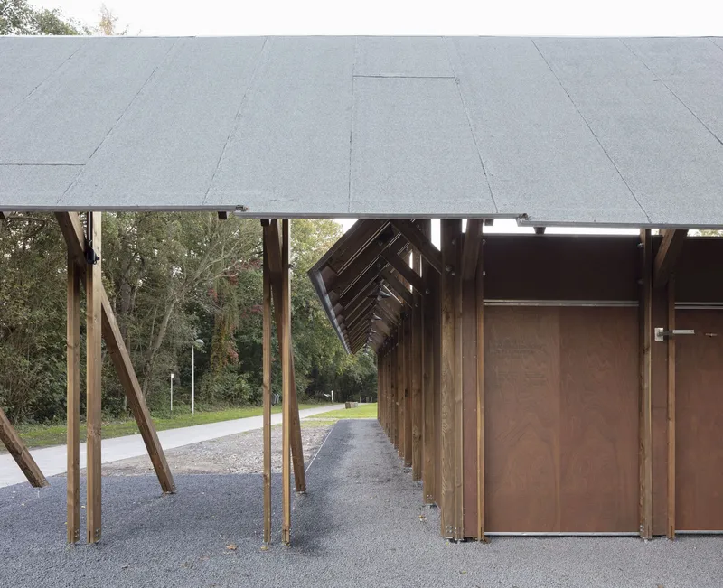 Covered walkway with angled timber columns and exposed roof structure above dark paneled doors