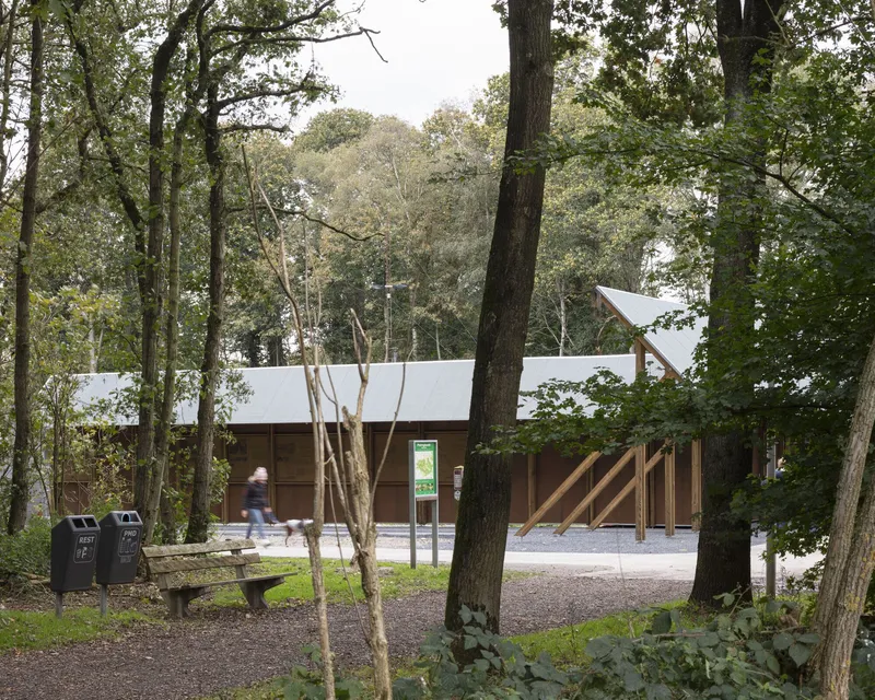 Timber pavilion with low-pitched metal roof set among mature trees with a walking figure
