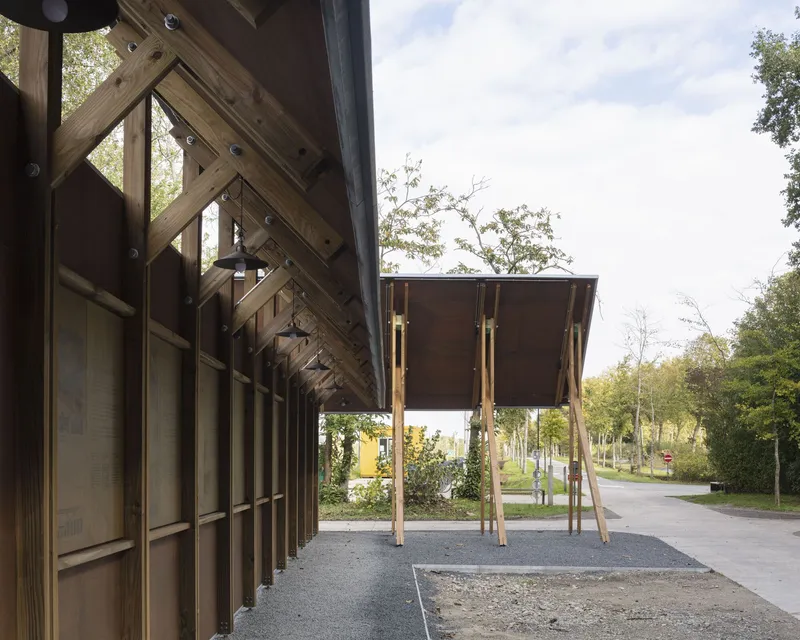 Timber canopy structure with slanted columns supporting a raised dark wood volume on stilts