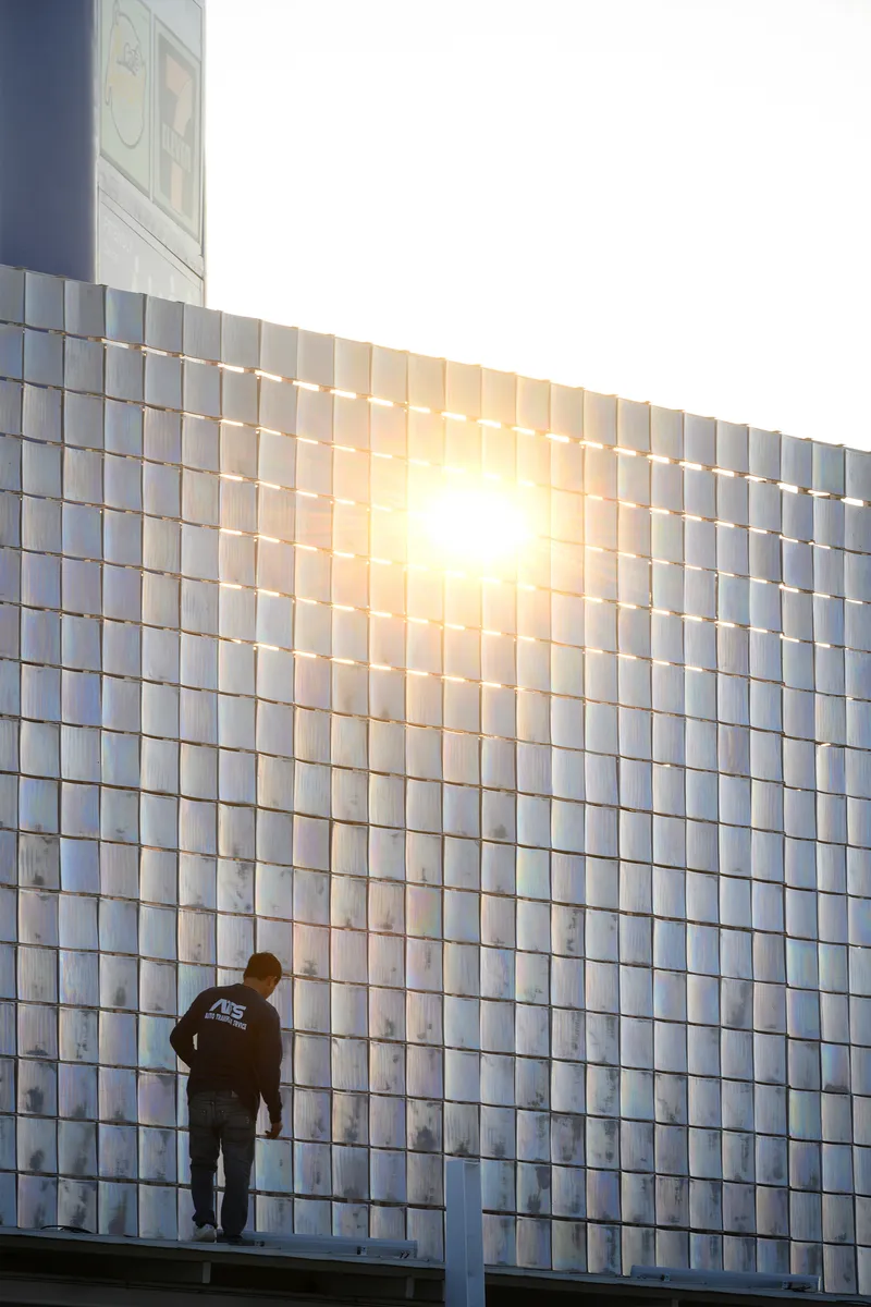 Figure standing on a rooftop platform beside the backlit glass tile facade with sun flare