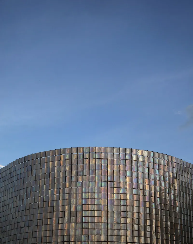 Curved facade clad in iridescent glass tiles catching late afternoon light against a clear sky