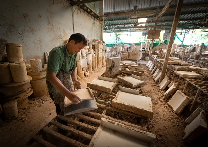 Worker placing ceramic tiles onto wooden drying racks beneath a corrugated metal roof