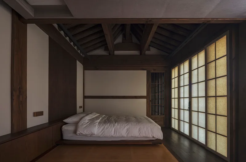 Bedroom with exposed roof trusses and backlit paper screens casting soft light at dusk