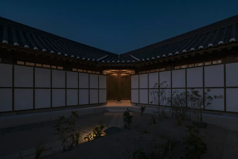 Interior courtyard with translucent wall panels and planted beds beneath converging tiled rooflines at twilight