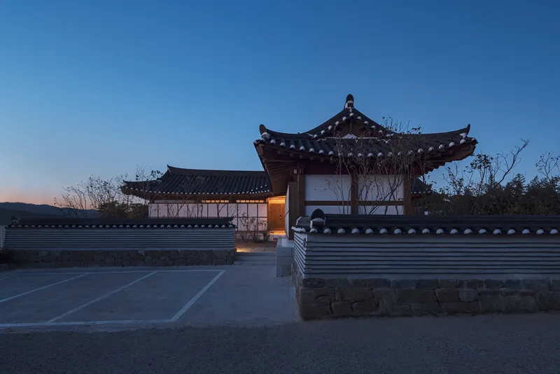 Entry gate and timber pavilions with curved tiled roofs framed by bare winter trees at sunset