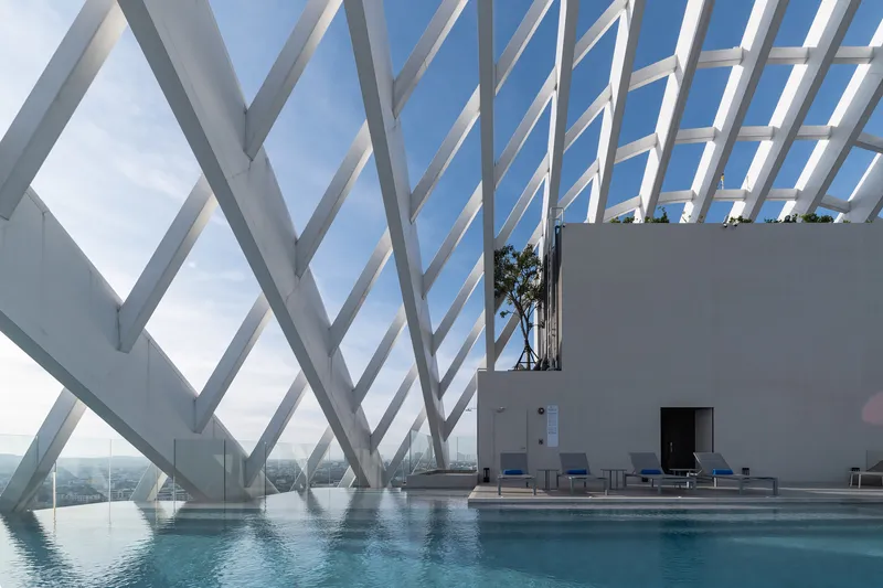 Rooftop infinity pool framed by white diagonal structural bracing against a clear blue sky