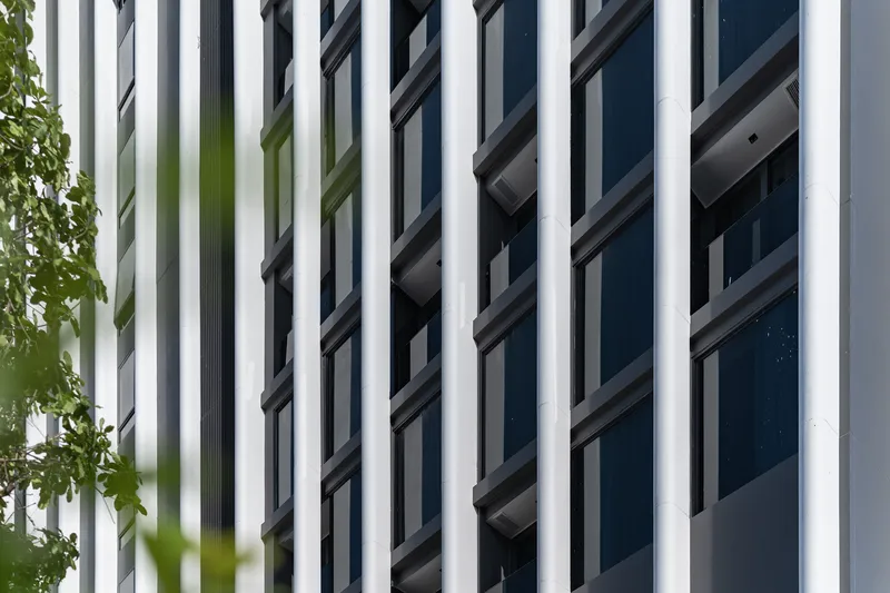 Vertical white fins alternating with dark glazed openings framed by green foliage at left
