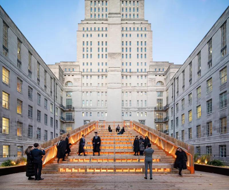 Illuminated timber and glass staircase bridging a courtyard between stone facades at dusk