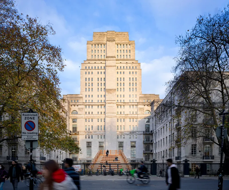 Stepped-back stone tower rising above the timber installation with pedestrians passing in the foreground