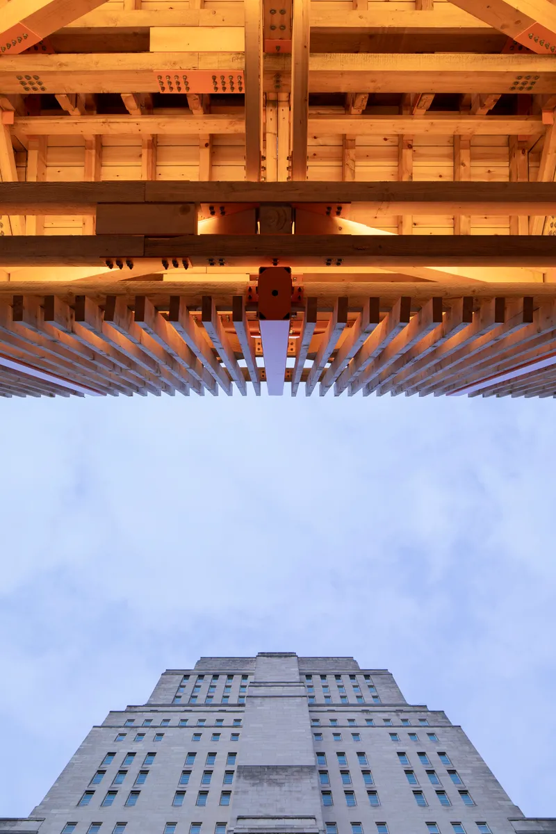 Upward view through layered timber joists and beams with a stepped concrete tower beyond