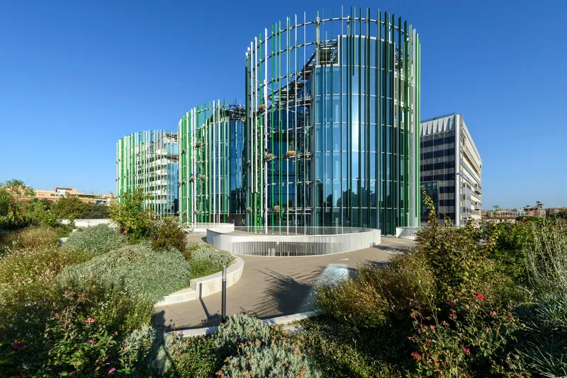 Glass tower wrapped in vertical green and white fins rising from a landscaped garden terrace under clear skies