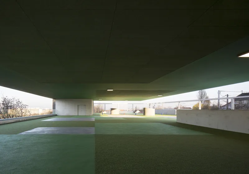 View beneath the green cantilevered ceiling with concrete columns and artificial lawn at dusk