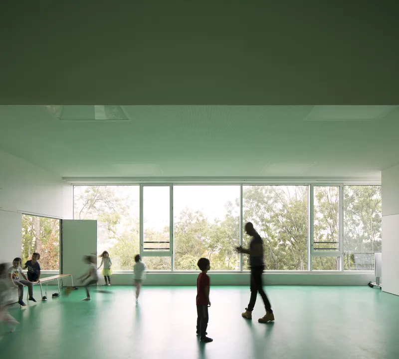 Interior classroom with green flooring and full-height windows as children move through diffused daylight