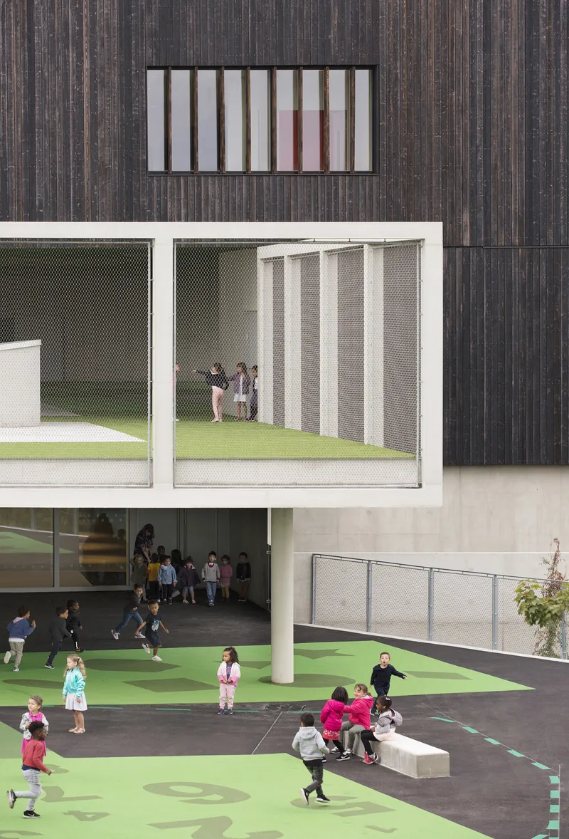Courtyard with green play surface, mesh-screened balcony, and children gathering under cantilevered volumes