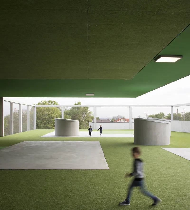 Covered play area with green soffit, cylindrical concrete elements, and children on artificial turf