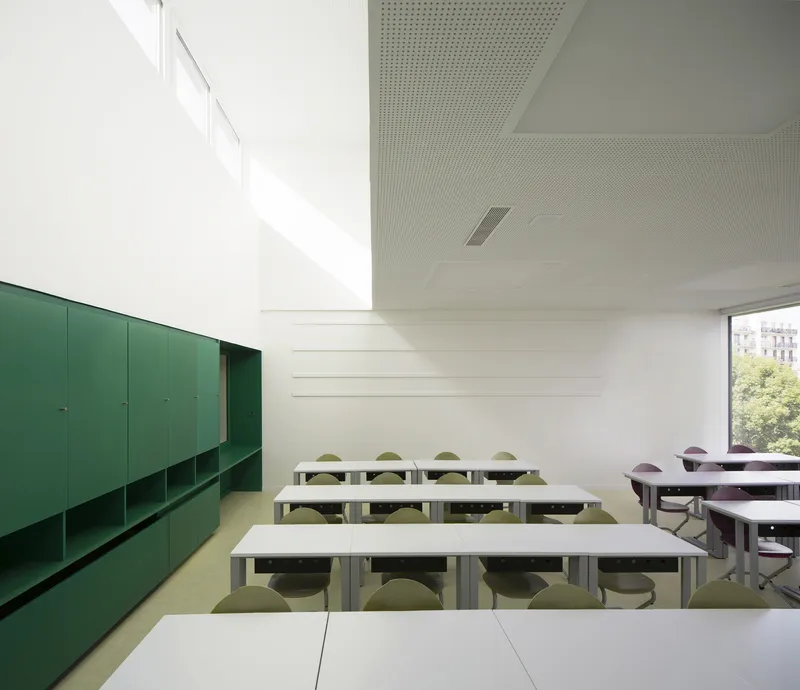 Classroom interior with green storage wall, white tables, perforated ceiling, and daylight from clerestory