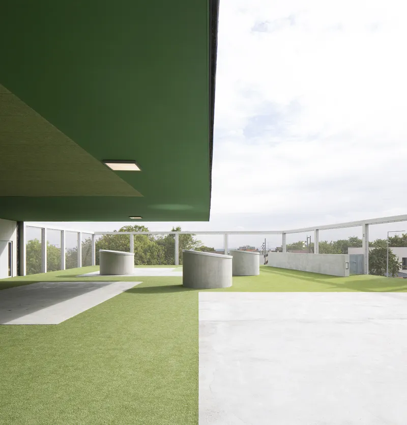 Covered terrace with green soffit and cylindrical concrete volumes on artificial turf under overcast skies