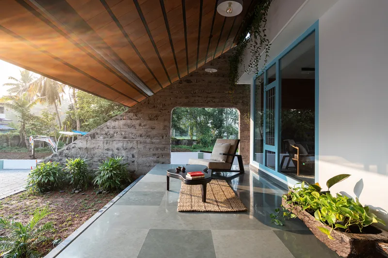Covered porch with exposed wood ceiling, laterite stone wall and planted beds flanking glass doors