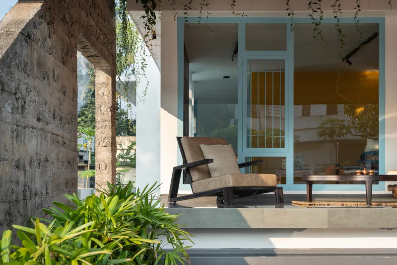 Concrete platform with lounge chair beside blue-framed glazing and laterite column under dappled shade