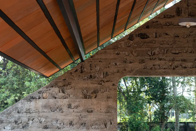Detail of laterite stone wall meeting timber ceiling with exposed black steel rafters and foliage beyond