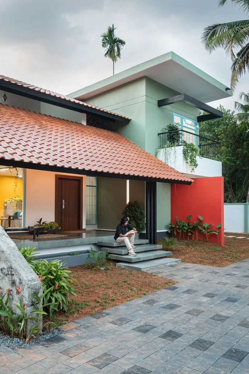 Street facade showing terracotta roof, coral red wall, and resident on entrance steps under palms