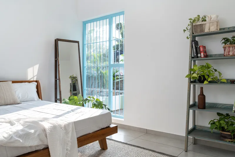 Bedroom with blue-framed door opening to a small balcony, potted plants and sunlight on white bedding