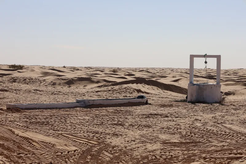 Concrete well structure with pulley system and adjacent water trough surrounded by tire tracks in sand