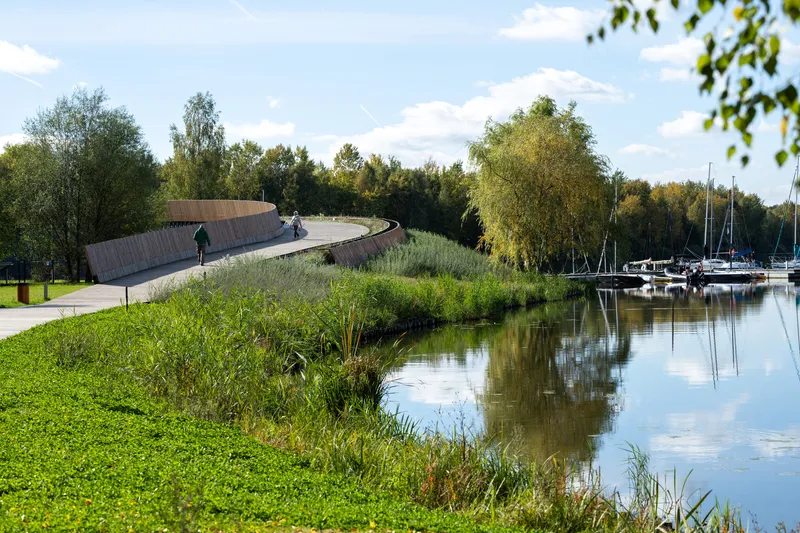 The elevated promenade curving past waterside planting beds with sailboats moored beyond on a sunny afternoon