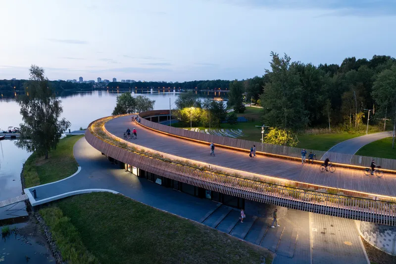 Evening view of the illuminated timber bridge with pedestrians and cyclists crossing above the waterfront plaza