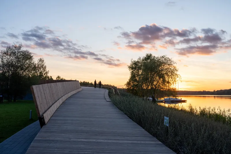 Wooden boardwalk with timber railing curving toward a lake at sunset with two silhouetted figures
