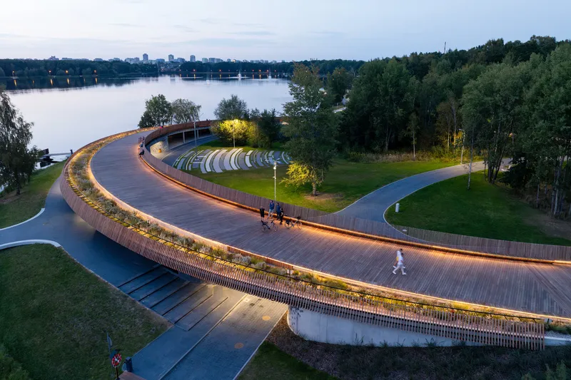 Elevated pedestrian boardwalk with integrated lighting and planted edge curving through the lakeside park at twilight