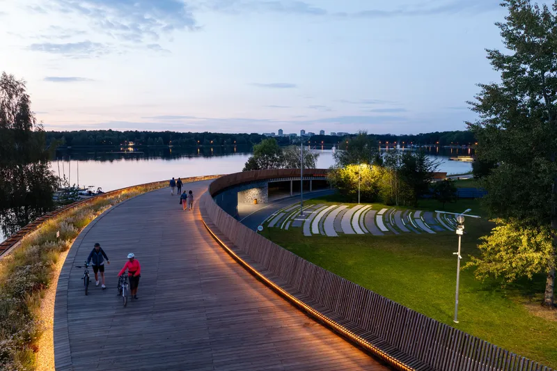 Cyclists and pedestrians on the timber boardwalk with amphitheater seating and illuminated tree at dusk