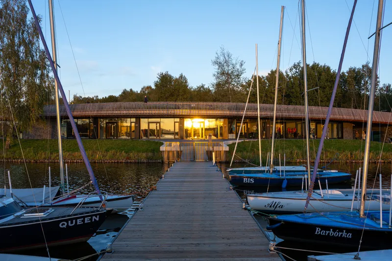 View from the timber dock through moored sailboats toward the glazed pavilion at sunset