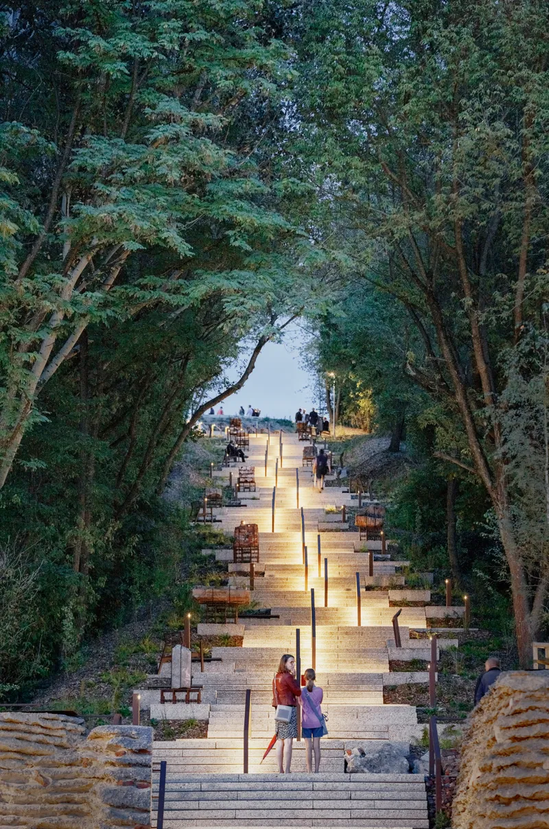 Illuminated stone stairway ascending through a canopy of trees at dusk with visitors scattered along the path