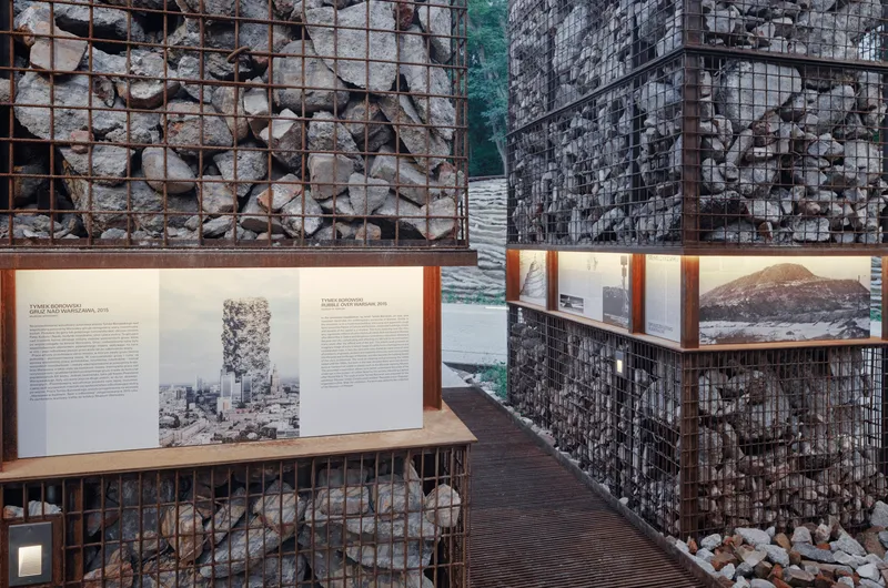 Exhibition wall of gabion baskets filled with stone supporting illuminated display panels and archival photographs