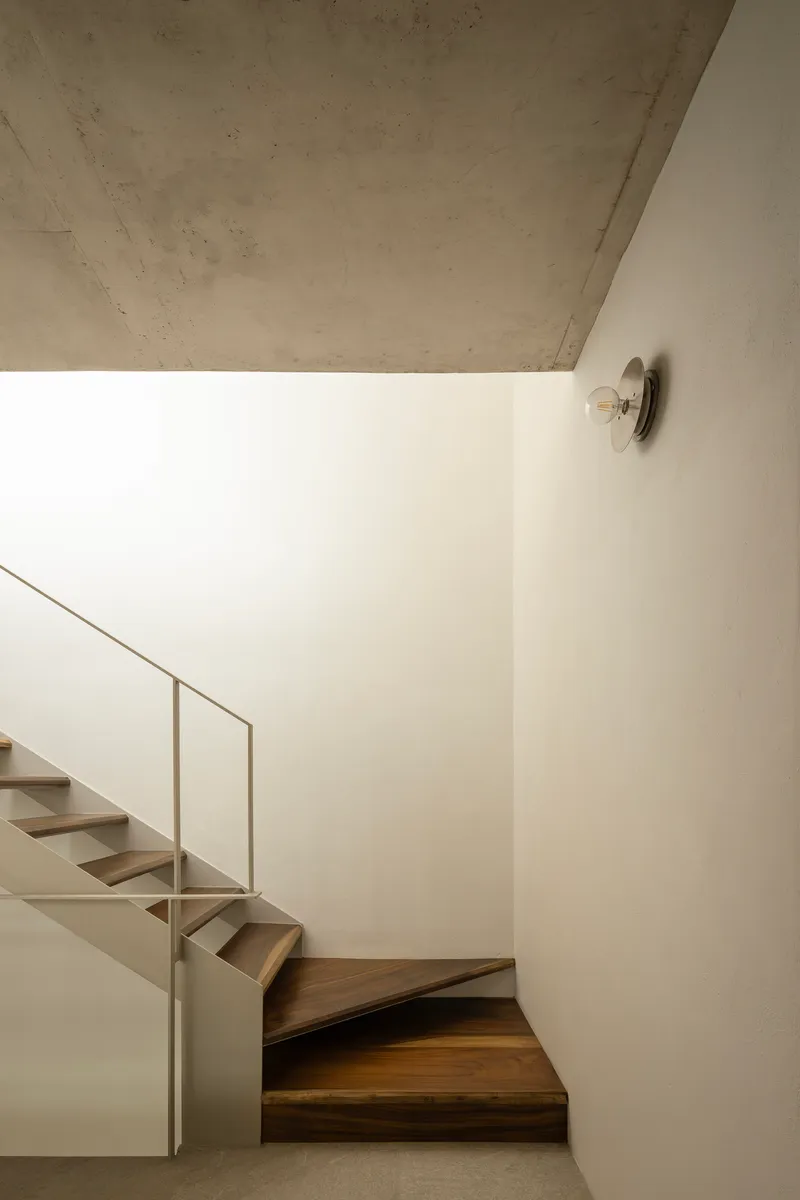 Wood staircase turning upward along white wall with circular sconce and concrete soffit above
