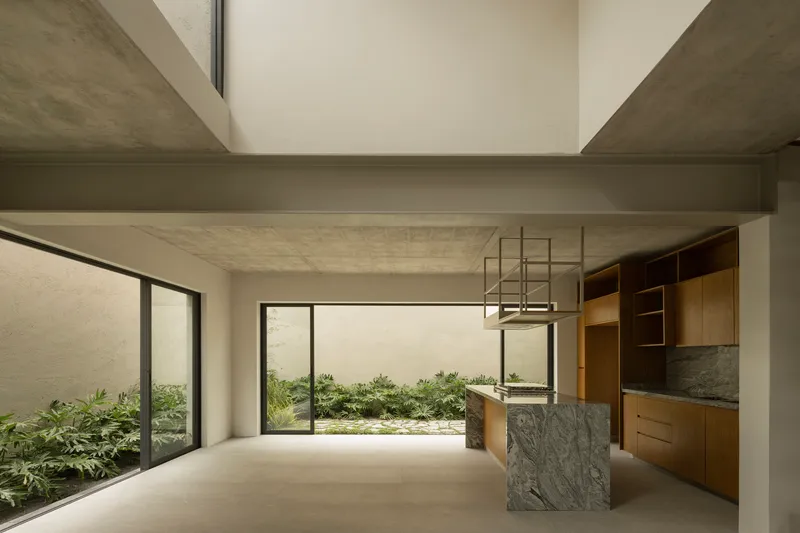 Open-plan kitchen and dining space with exposed concrete ceiling beam framing a view to planted courtyards