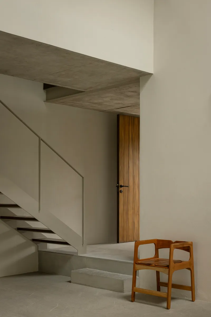 Concrete staircase rising alongside a timber door and wooden chair in a double-height interior volume