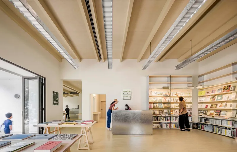 Reception area with metal desk and plywood bookshelves beneath exposed ceiling beams and linear lighting