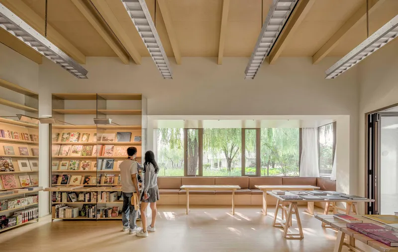 Timber ceiling beams and recessed light slots above a bookshop reading room with visitors browsing near the windows
