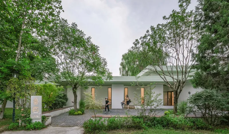 Front facade with vertical window openings and brick paving framed by mature trees under an overcast sky