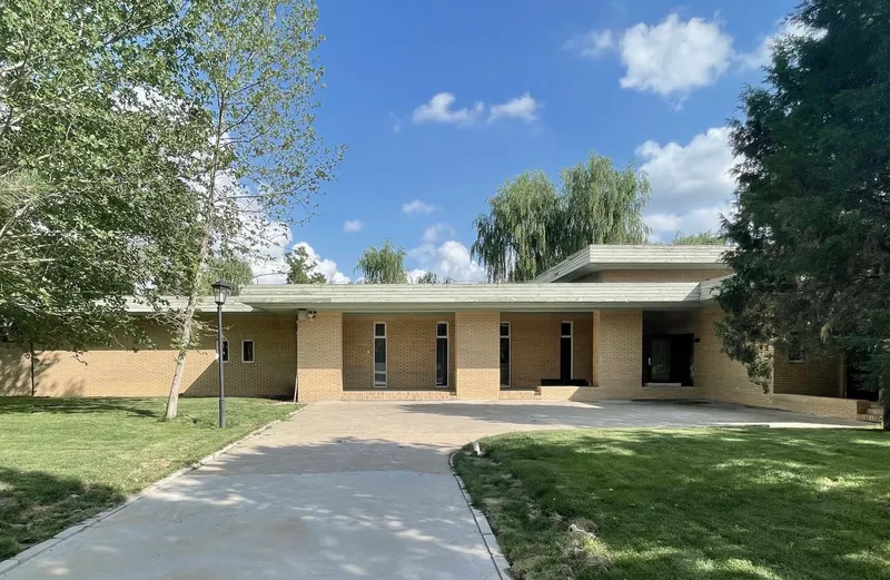 Front facade of buff brick with flat roof and tall willow trees framing the lawn